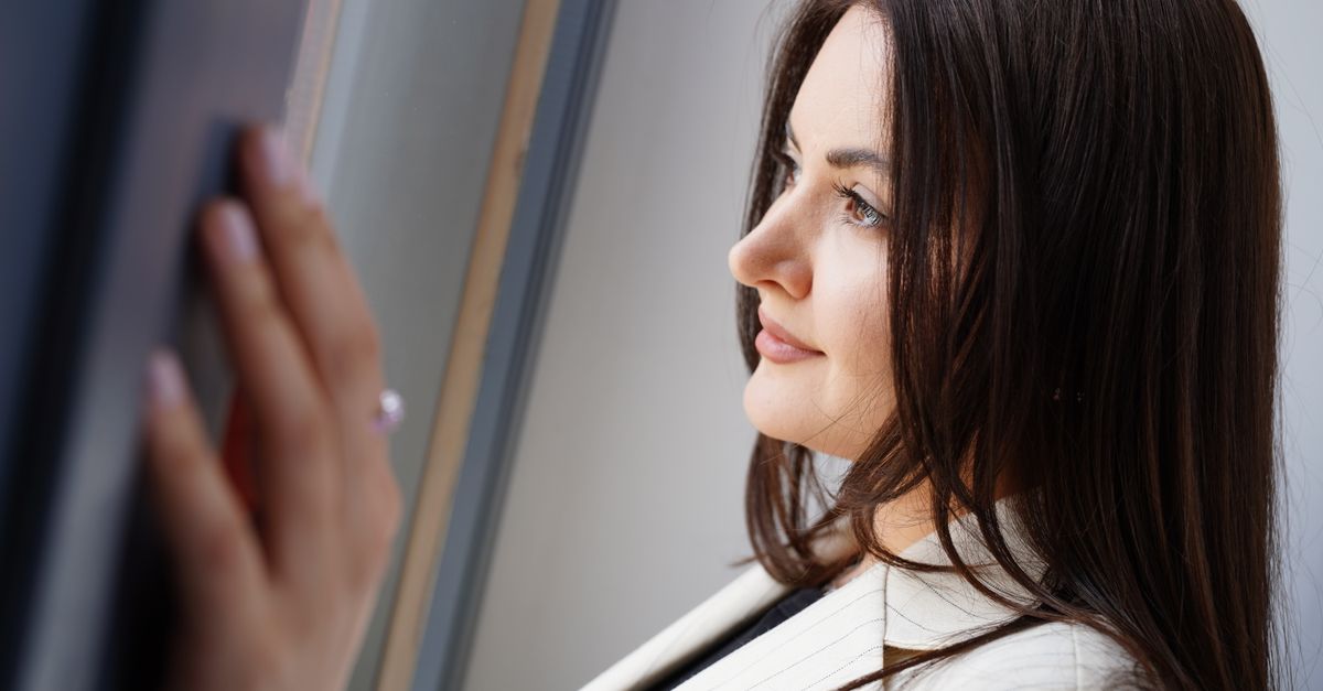 A thoughtful woman in professional attire gazing through a window indoors.
